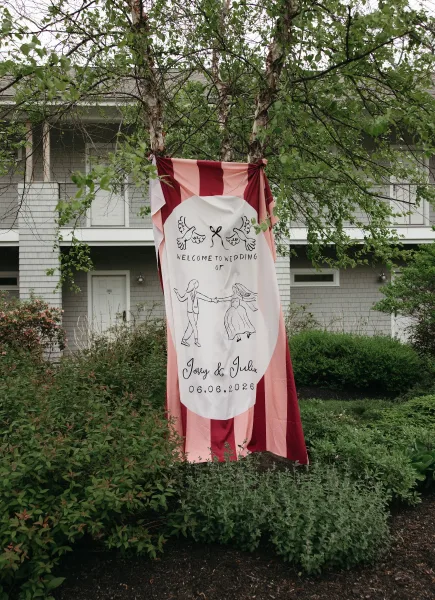 Wedding welcome sign, a fabric wedding sign featuring a dove illustration, displayed outdoors amid trees, shrubs, and a house exterior.