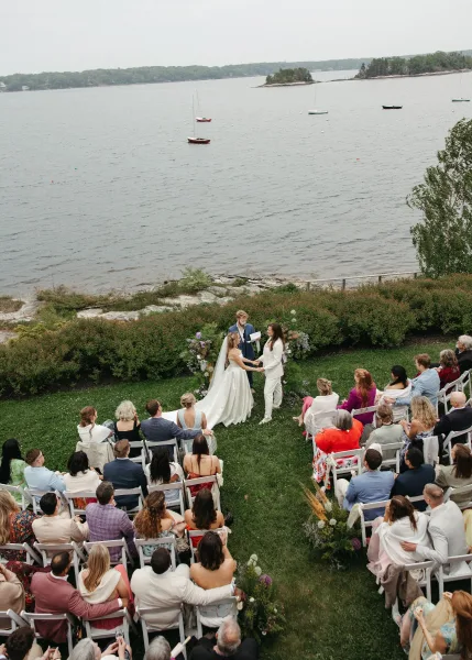 Ceremony moment with the couple exchanging vows outdoors on a waterfront lawn with sailboats and small islands, the bride’s long veil visible.