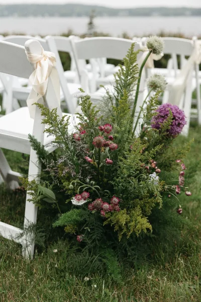 Ceremony aisle flowers form an airy aisle floral arrangement flanking white folding chairs on a grass lawn by the water and distant shoreline