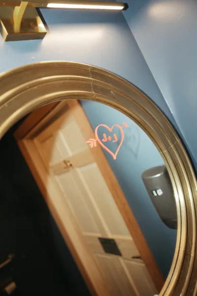 Wedding mirror sign with a bathroom mirror message and a pink heart doodle, set on a blue wall near a wooden door and soap dispenser.