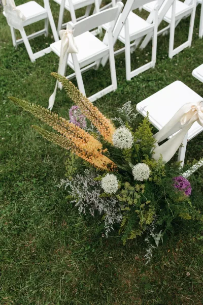 Ceremony aisle flowers create a lush aisle floral arrangement flanking white folding chairs, elegantly framing the path.