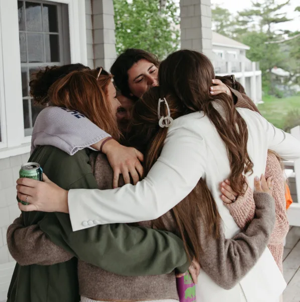 Bridal party hug as bridesmaids with bride in a white blazer squeeze close on a porch, a tender getting ready moment before the ceremony.