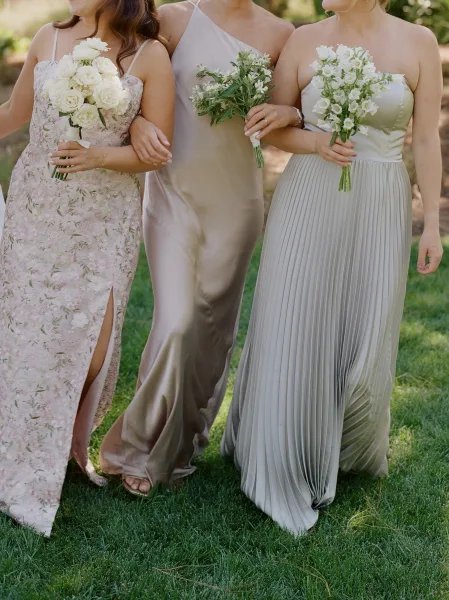 Bridesmaid dresses in mismatched neutral bridesmaid dresses, each holding white rose bouquets, standing on a green lawn with trees behind
