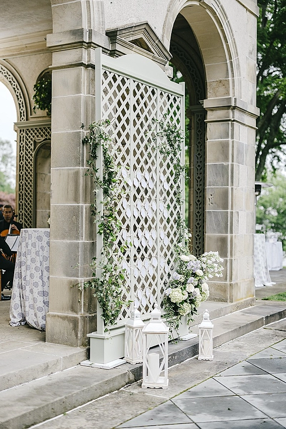 Wedding ceremony backdrop with a white lattice screen draped in greenery garland, white florals and lanterns under stone pavilion arches