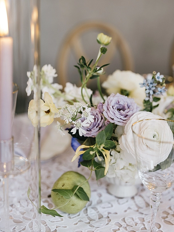 Wedding centerpiece with pastel wedding centerpiece blooms of white ranunculus and lavender roses, greenery, and a tall taper candle on lace tablecloth