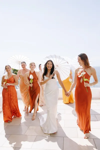 Bride with bridesmaids walking on a coastal terrace, carrying bouquets and white parasols, ocean and palm trees behind them