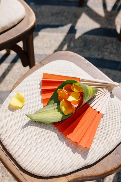 Ceremony chair detail with wedding ceremony fans and a calla lily on a cushion, rose petals scattered on stone pavement in sunlight