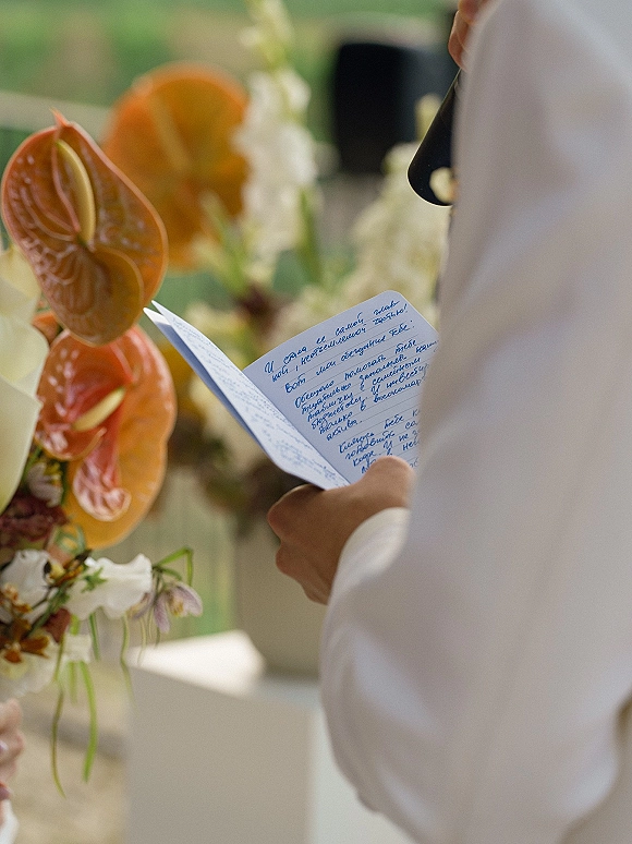 Wedding vows in a handwritten wedding vows booklet held near a boutonniere, with anthurium and white blooms against blurred outdoor greenery