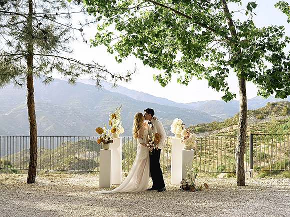 Wedding kiss outdoors as the bride holds a bouquet and veil drapes behind, framed by floral pedestals with mountain views beyond