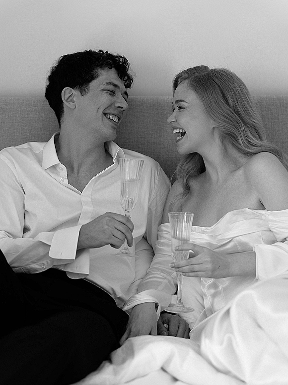 Couple portrait in black and white wedding portrait style, bride and groom laughing on an upholstered sofa, toasting champagne flutes by a plain wall