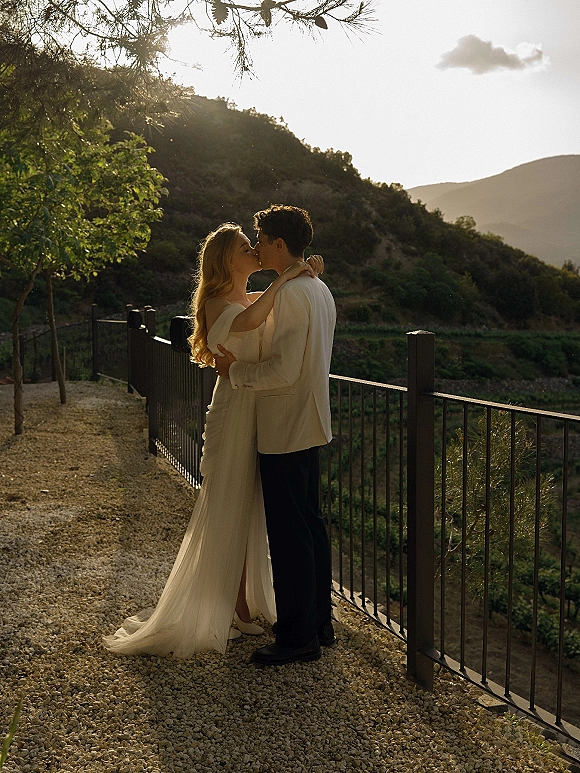 Wedding kiss portrait of bride in off the shoulder dress and long veil embracing groom at sunset overlook with mountain landscape behind