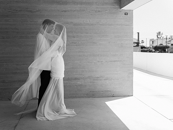 Wedding couple portrait of bride and groom under veil, foreheads touching in bright sunlight on an outdoor terrace by a concrete wall