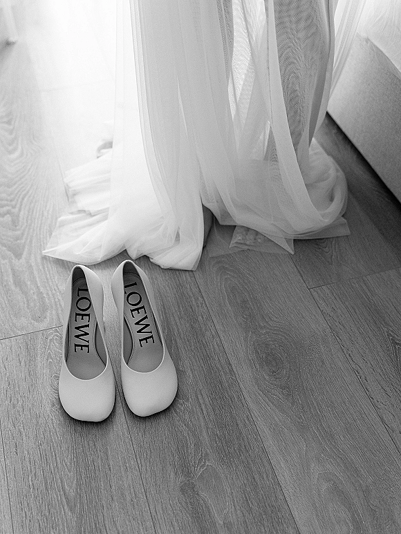 Bridal shoes arranged in a wedding shoes flat lay with white heels beside a veil and dress hem on a wood floor in window light