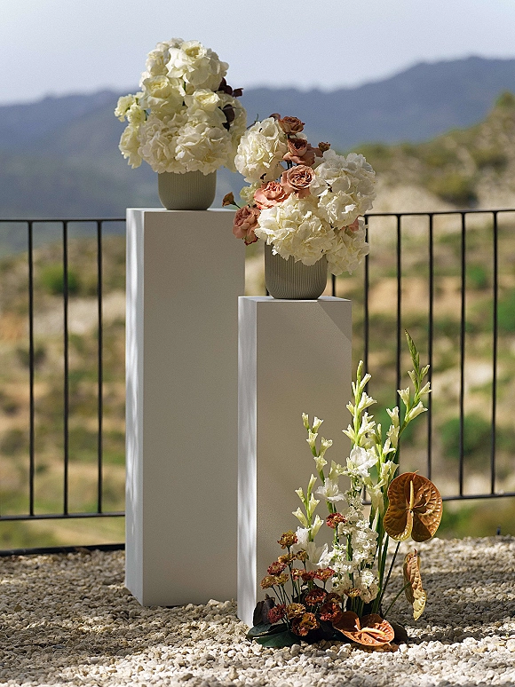 Wedding floral arrangement with white hydrangeas and blush roses in ceramic vases on pedestals, set against a mountain landscape backdrop