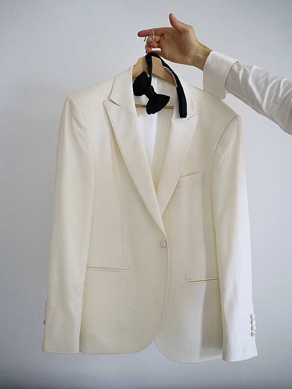 Groom suit, an ivory tuxedo jacket with black bow tie on a wooden hanger, held up against a clean white wall background