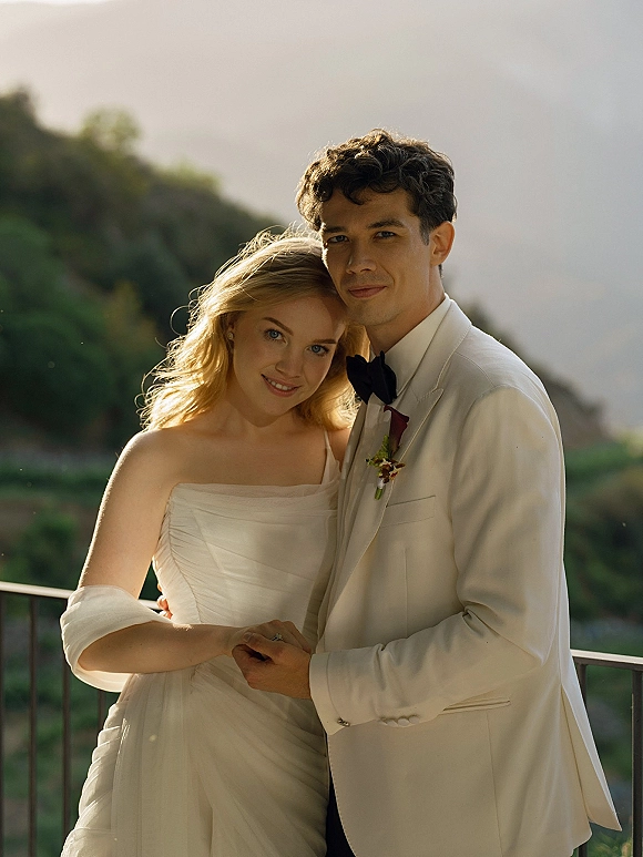 Couple portrait of bride and groom embracing, her off-shoulder wrap and ring visible, on a balcony with hills and sky behind