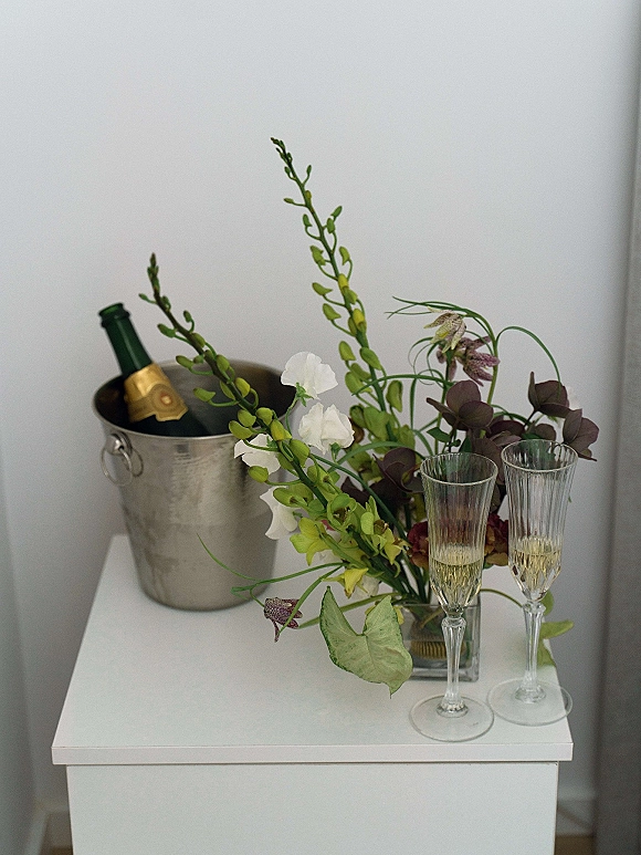 Champagne toast setup with champagne flutes on a side table, champagne bottle in a metal ice bucket beside flowers against a white wall