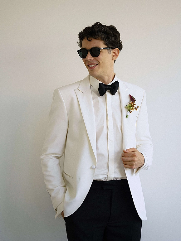 Groom portrait in a white tuxedo groom look, wearing black bow tie and sunglasses with a boutonniere against a neutral wall