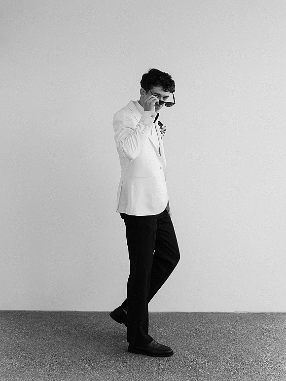 Groom portrait in a white tuxedo jacket with black bow tie and boutonniere, wearing sunglasses, standing against a plain wall.