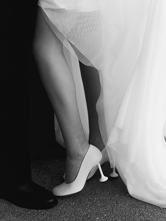 Wedding shoes with white bridal heels beside the groom’s dress shoes, framed by gown hem and veil on a carpeted floor