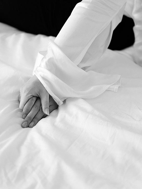 Couple holding hands, bride and groom hands intertwined on white bed sheets, long-sleeve bridal robe and manicure in view
