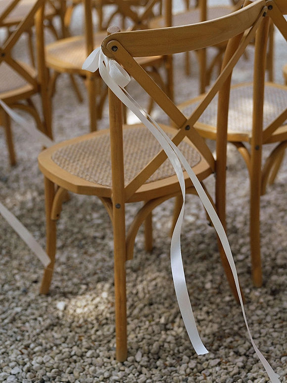 Ceremony chairs with white ribbon ties on wooden crossback wedding chairs arranged in neat rows over gravel ground outdoors