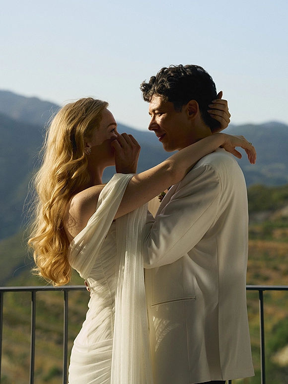 Couple portrait of bride and groom embrace with a forehead touch, bride in draped-sleeve gown on a balcony with mountain view behind