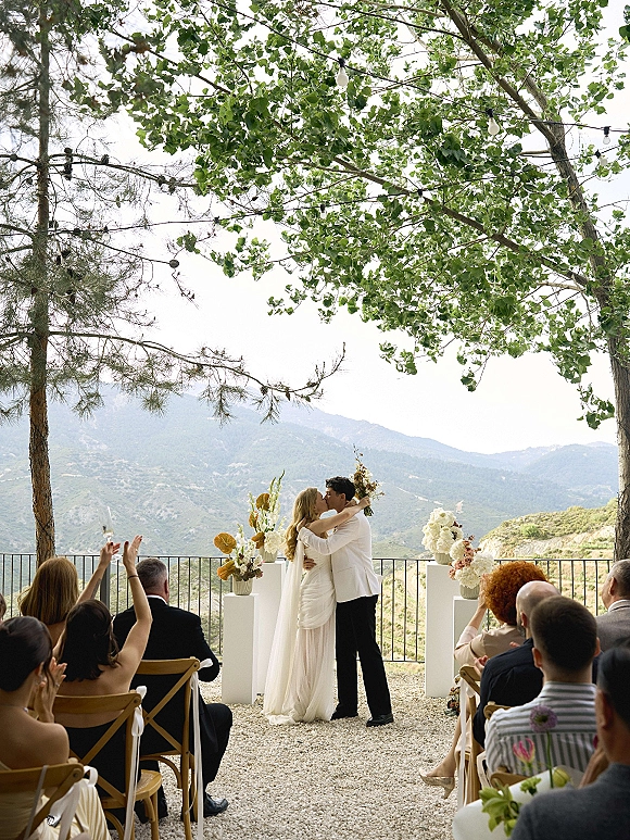 Ceremony kiss as bride in veil and groom in suit embrace beside floral pedestals on a terrace with mountain view and string lights