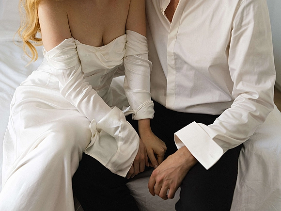 Couple portrait of bride and groom on bed, holding hands on white linens; bride in strapless satin gown with opera gloves, groom in white shirt