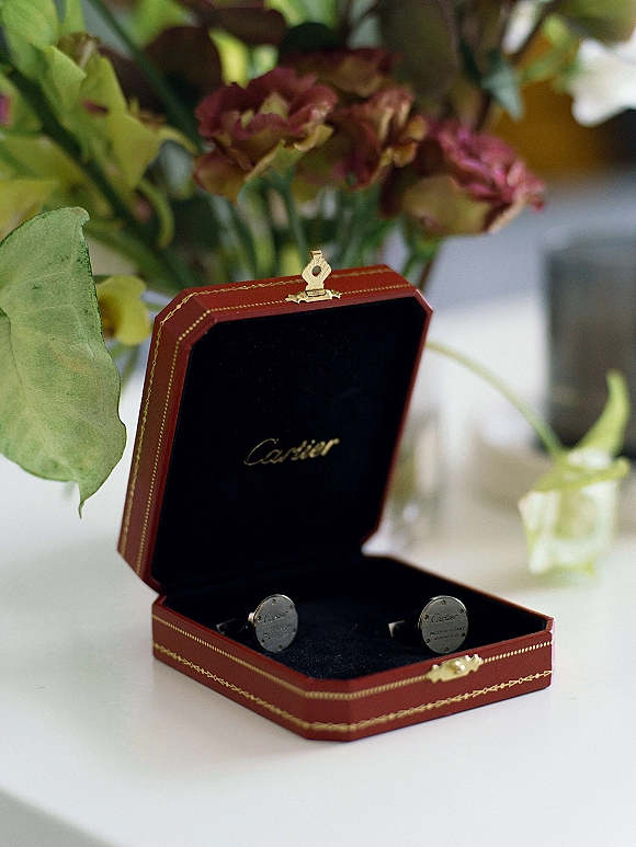 Wedding cufflinks displayed in a red jewelry box on a white tabletop with soft flowers and greenery accents nearby