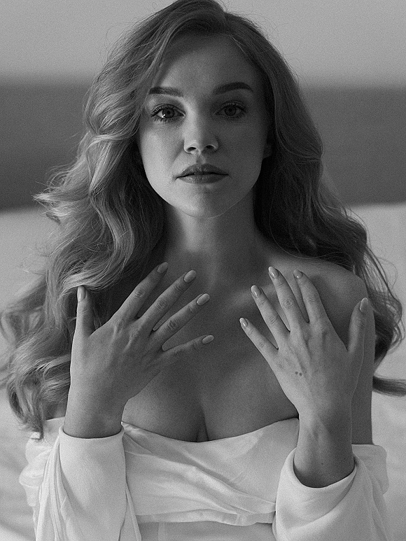 Bridal portrait of a bride with long wavy hair and soft glam makeup in an off-shoulder gown, reclining on a bed in a bedroom interior