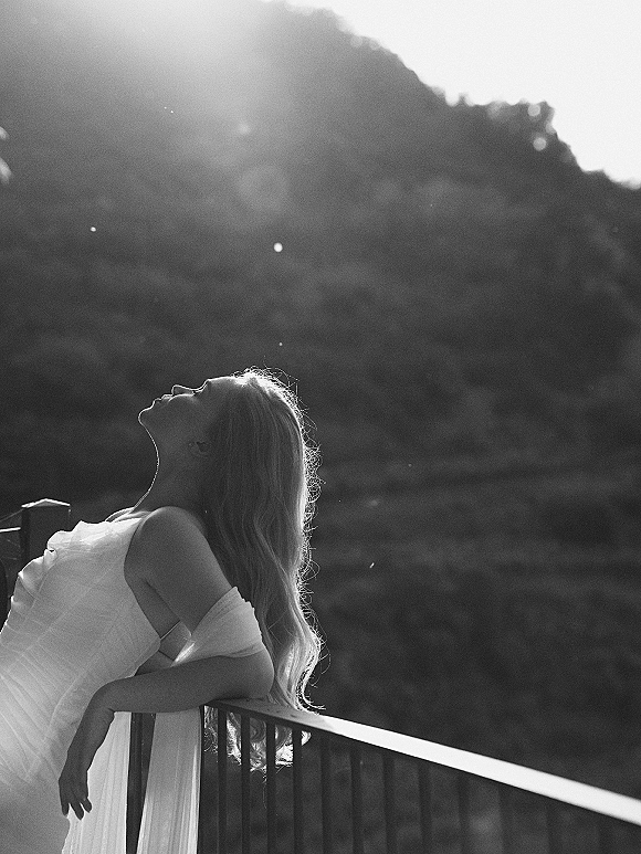 Bridal portrait of a bride in an off-the-shoulder wedding dress leaning on a balcony railing, backlit by sun flare over hills