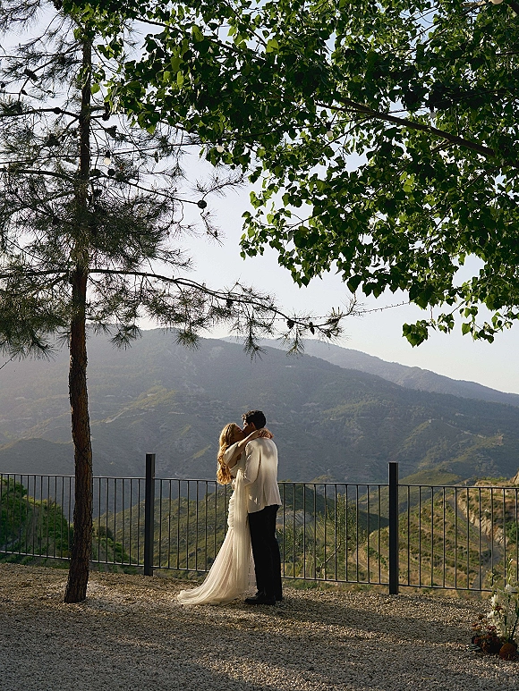 Wedding couple portrait of bride and groom embrace under string lights by a metal railing, with mountain view and trees behind them