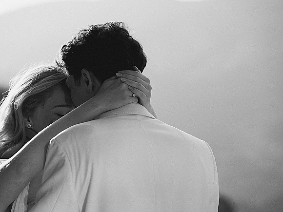 Wedding kiss in a black and white wedding photo, close up as the bride cradles the groom’s face, engagement ring visible by a plain wall