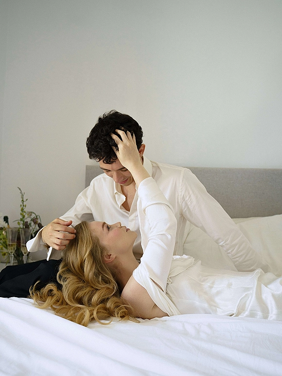 Couple portrait of bride and groom in bed, cuddling on white linens with champagne flutes beside them in a neutral bedroom setting