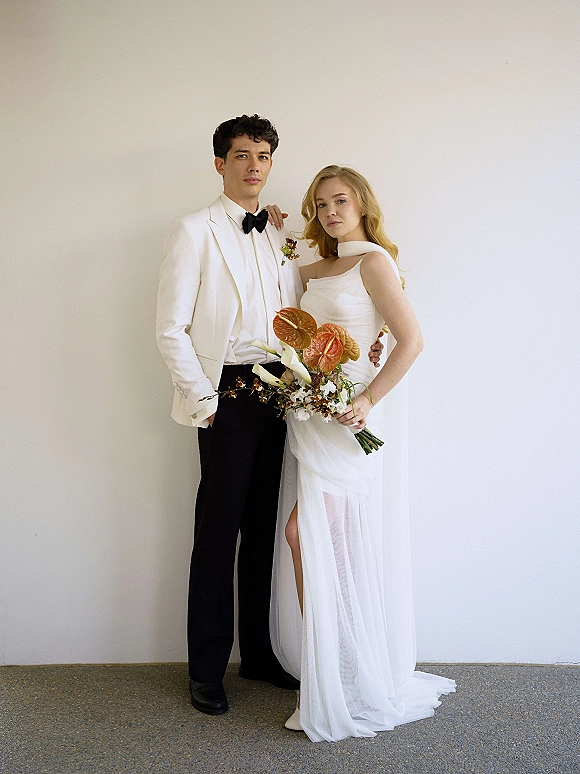 Couple portrait of bride in a high-slit gown with cape and groom in white tuxedo, holding anthurium bouquet against a plain studio wall