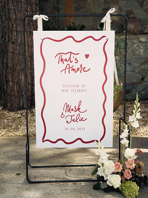 Wedding welcome sign with custom wedding welcome sign styling, red script typography and ribbon bows, roses and gladiolus by a stone wall