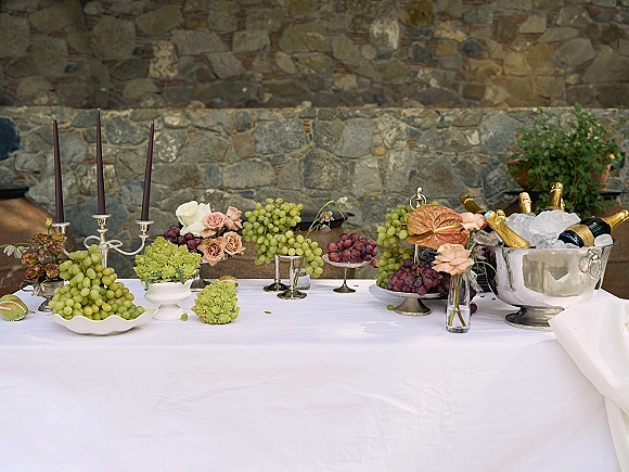 Champagne display table with a wedding champagne station of bottles in an ice bucket, candelabra, roses, and grapes on a patio by a stone wall