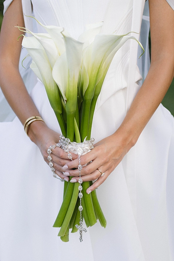 Calla lily bouquet of white blooms held in bridal hands with a crystal rosary and cross pendant, engagement ring, and soft greenery behind