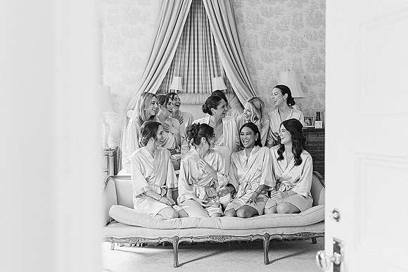 Bridesmaids getting ready in bridal party robes, laughing in satin robes on a bed in a patterned bedroom with soft lamps