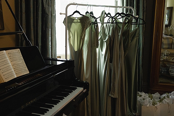 Bridesmaid dresses in sage green hang on a rack by a bright window, satin slips beside a piano with sheet music in a cozy living room