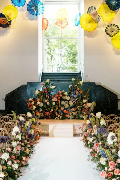 Ceremony aisle decor with a floral aisle runner bordering a white runner, leading to a platform beneath colorful glass wall sculptures indoors