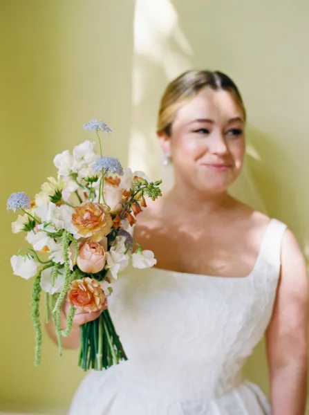Bridal portrait of a bride holding bouquet, wearing a white square-neck dress and pearl earrings in soft window light against a painted wall