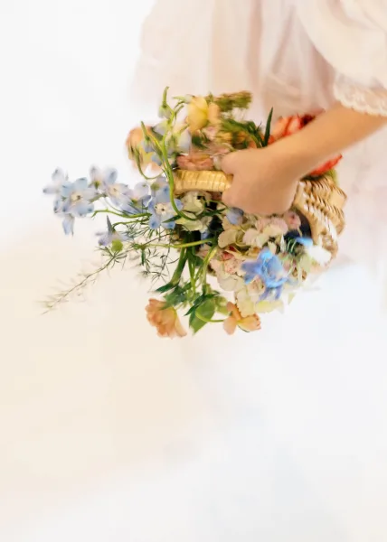 Bridal bouquet of wildflowers arranged in a wicker basket, with pastel blooms and greenery held by a white dress sleeve against a white backdrop