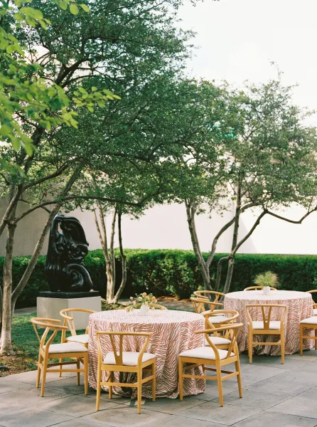 Reception tablescape with outdoor reception tables, patterned tablecloths, wooden chairs, and bud vase centerpieces on a garden patio near a sculpture