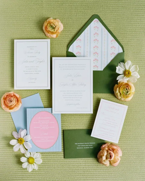Wedding invitation suite flat lay with green envelope, floral liner, RSVP and details cards, plus peach ranunculus on textured fabric backdrop