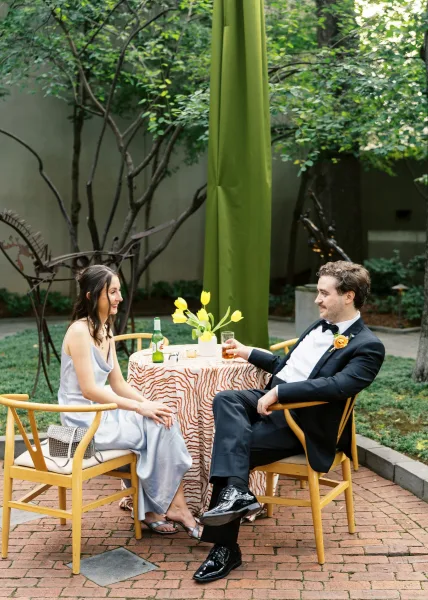Couple portrait of bride and groom seated at a round cocktail table with yellow tulip centerpiece in a garden courtyard on brick patio