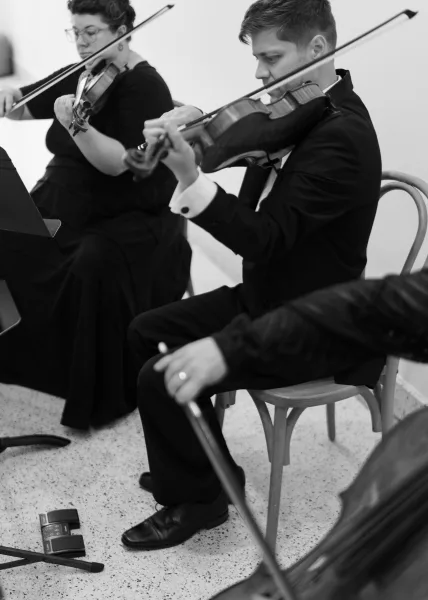 Wedding musicians in formalwear performing on violins with a sheet music stand and chairs against a simple indoor wall and floor