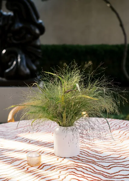 Wedding centerpiece with greenery table centerpiece grasses in a white ceramic vase on patterned tablecloth, votive candle, hedged patio backdrop