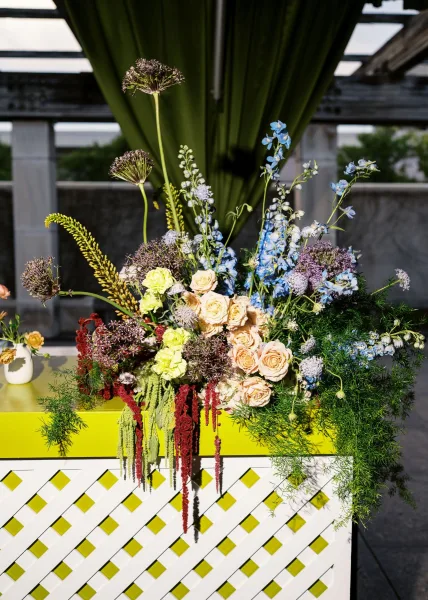 Wedding floral arrangement spilling garden roses and delphinium from a white vase on a patterned pedestal with green drapery on an outdoor patio